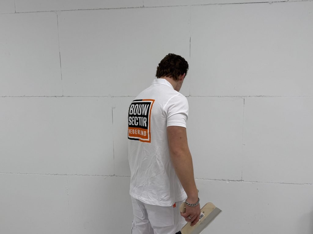 Stukadoor in wit werkpak bekijkt een kale wand van dichtbij, met de spaan in zijn hand