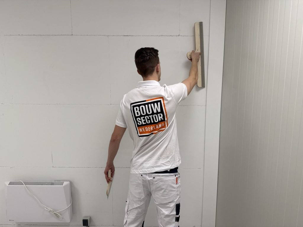 Stukadoor van Bouwsector Nederland houdt een lange spaan hoog tegen een witte wand, met een kachel op de vloer