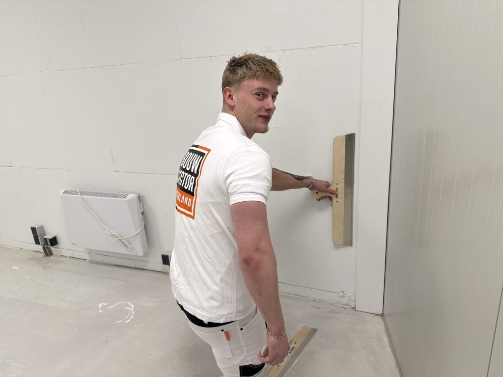 Jonge stukadoor kijkt om terwijl hij met een rei langs de onderkant van een wand gaat