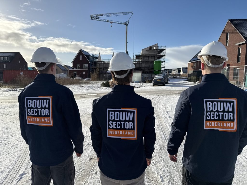 Drie vakmensen van Bouwsector Nederland met witte helm op een besneeuwd terrein met nieuwbouwwoningen en een bouwkraan